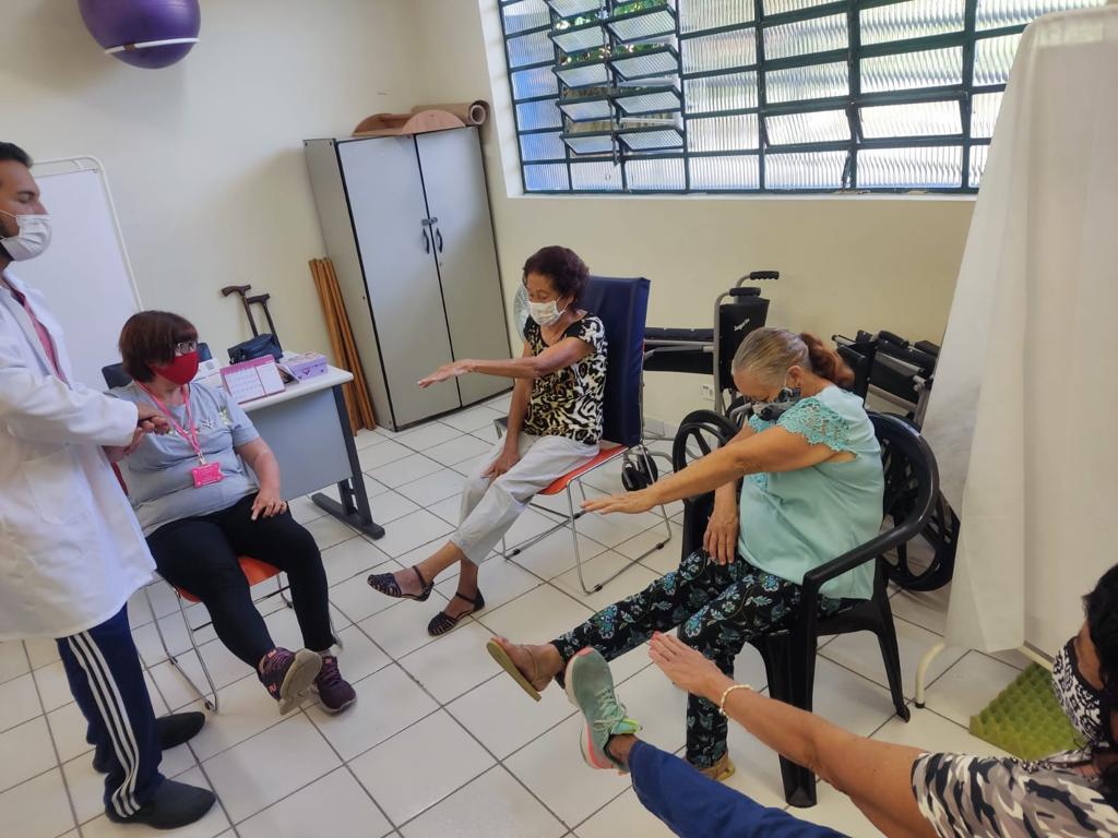 A foto mostra um grupo de mulheres idosas sentadas em cadeiras. Elas estão com a pernas direitas elevadas e esticadas e os braços esquerdos de cada uma também estão esticados. Elas são acompanhadas e recebem orientações de um profissional da saúde, homeme branco que está de pé, ao lado das mulheres.
